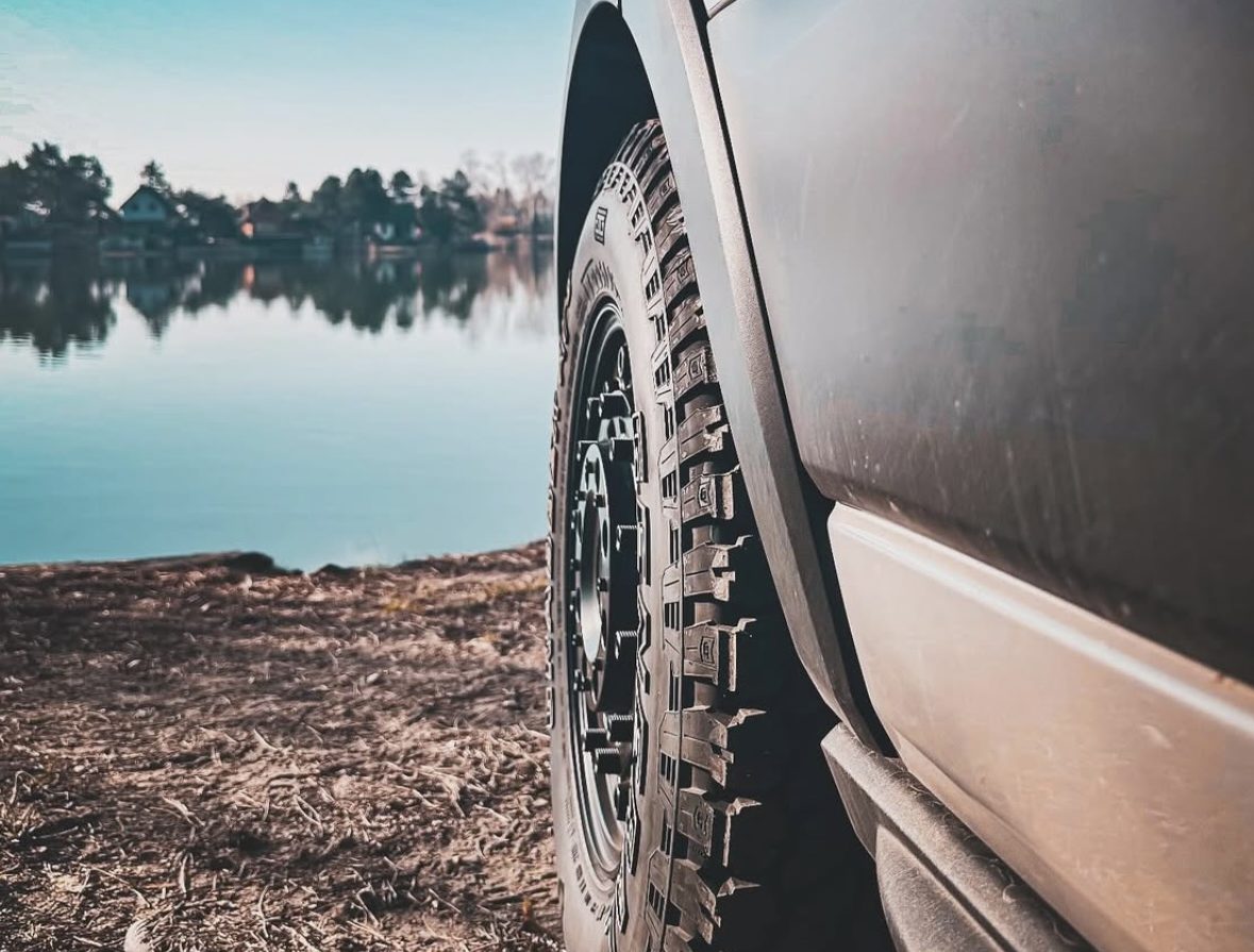 An all-terrain tyre at ground level, standing out from the arch of a grey van, a lake behind