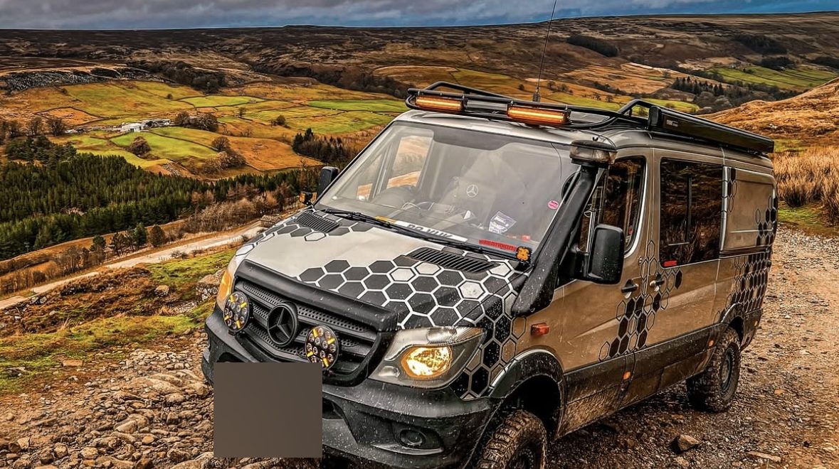 A 4x4 Mercedes-Benz Sprinter with a hex wrap and roof lights on a rocky moor track