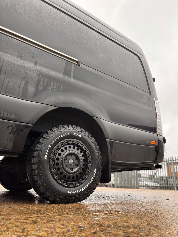 A matte black beadlock-style Rogue wheel on a panel van conversion in a rainy industrial yard