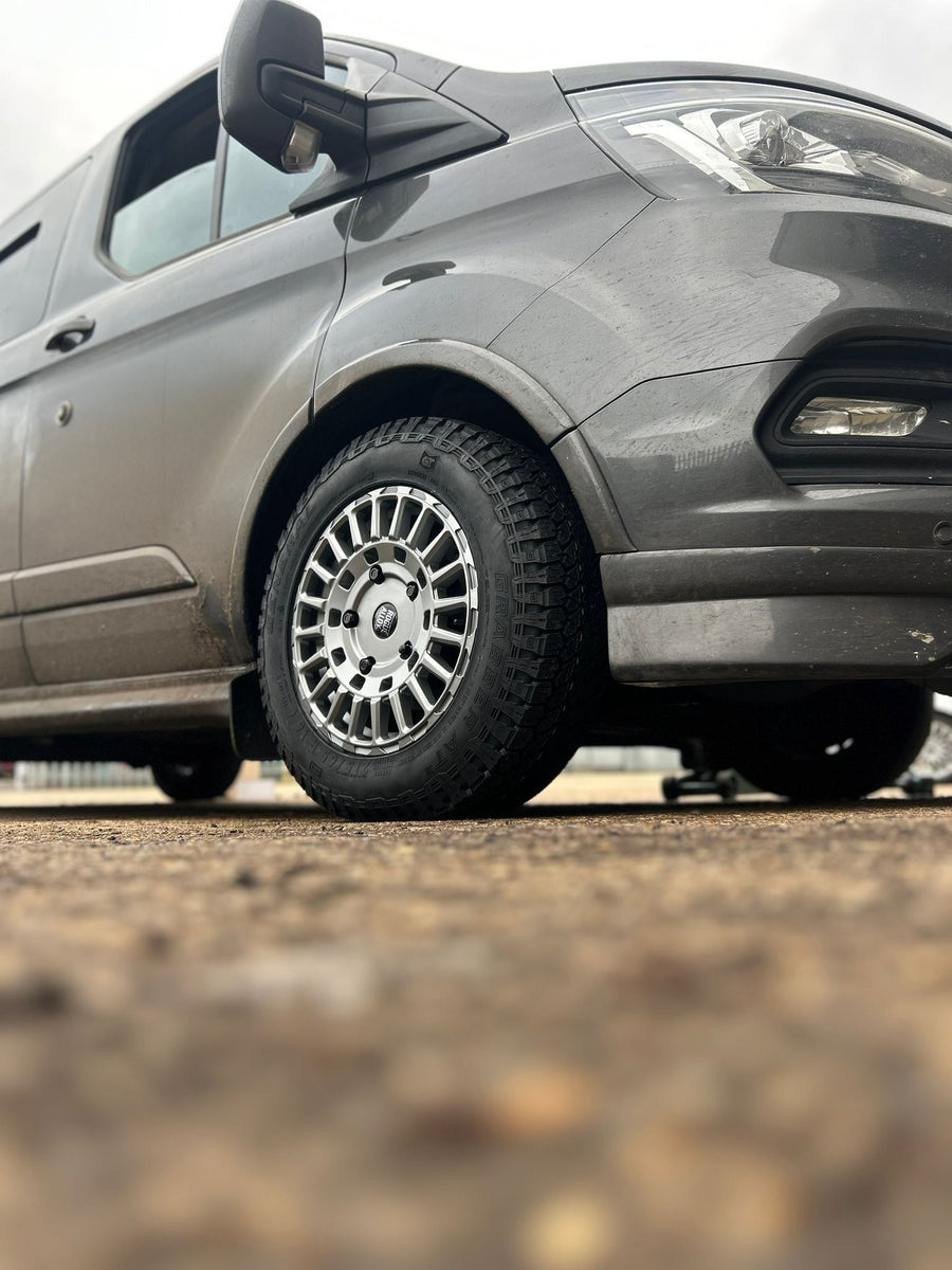 A bright satin-silver multi-spoke Rogue wheel on a Ford Transit Custom, General Grabber AT3, in a muddy yard